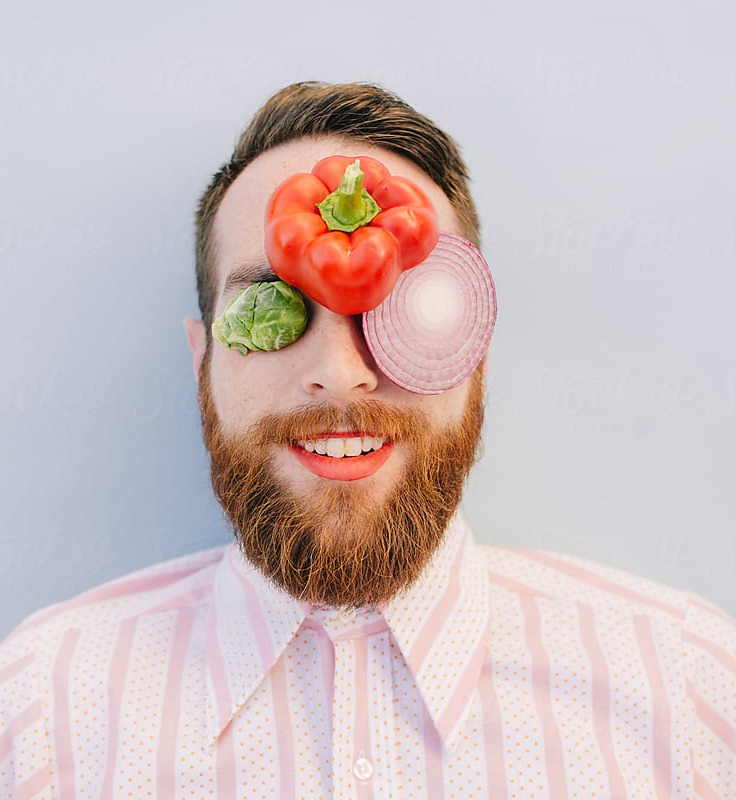 A smiling bearded man, vegetables held in front of one eye